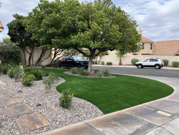 Residential corner lot with curved artificial grass lawn, decorative rock landscaping, and mature shade trees for low-maintenance curb appeal.