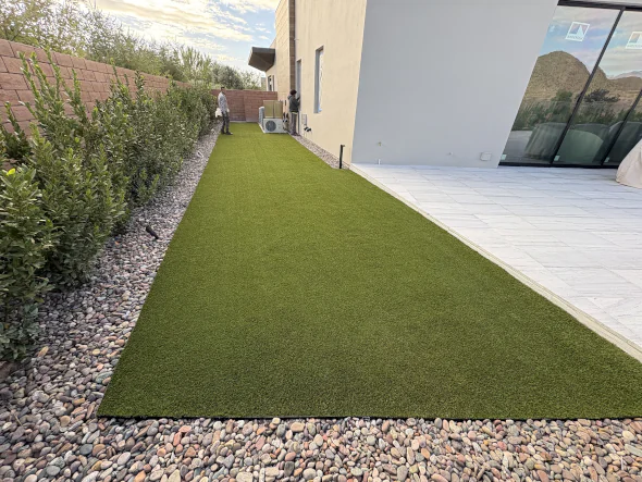 Artificial grass installed in a modern backyard alongside a house, featuring a clean stone border and landscaped greenery for a low-maintenance outdoor space.