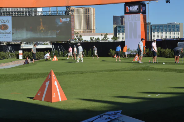 Event golf setup featuring large-scale artificial turf installation with smooth putting greens, practice stations, and players participating in a professional outdoor tournament environment.