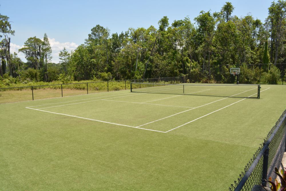 Artificial turf tennis court installed by Southwest Greens, with a basketball hoop visible beyond the net and mature trees surrounding the property.