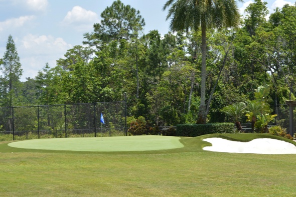 Wide view of a Southwest Greens synthetic putting green with sand bunker, palm trees, and a covered outdoor seating pavilion in the background.