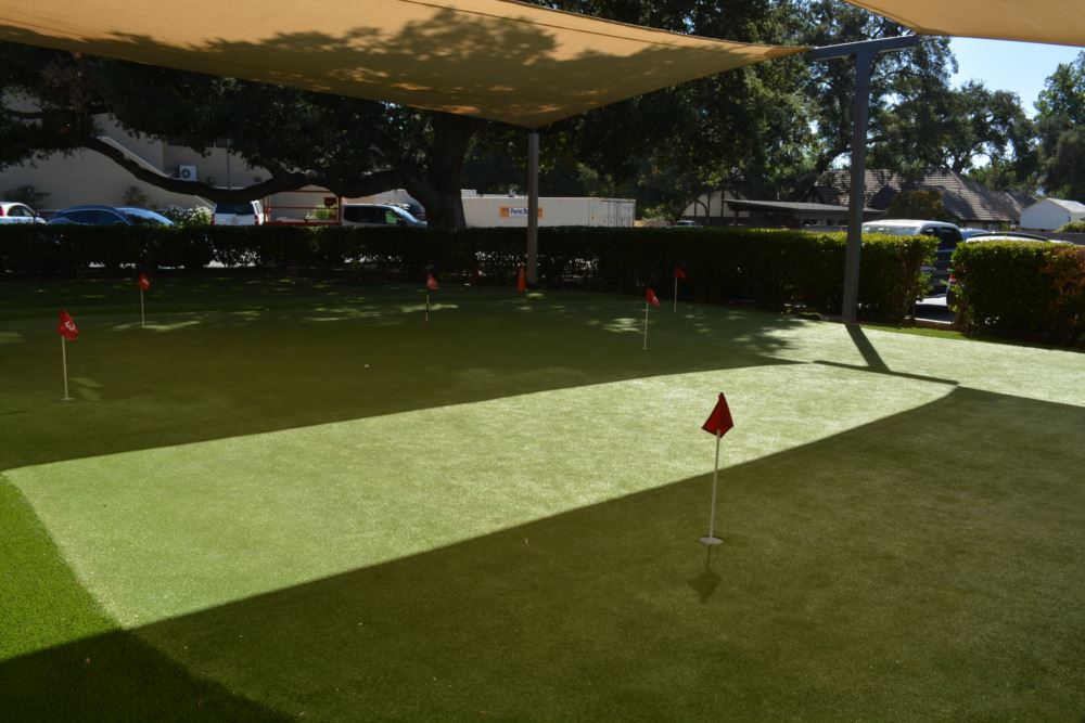 Artificial putting green with tee line zones, red flag pins, and a shade sail canopy at a commercial golf practice facility surrounded by mature oak trees