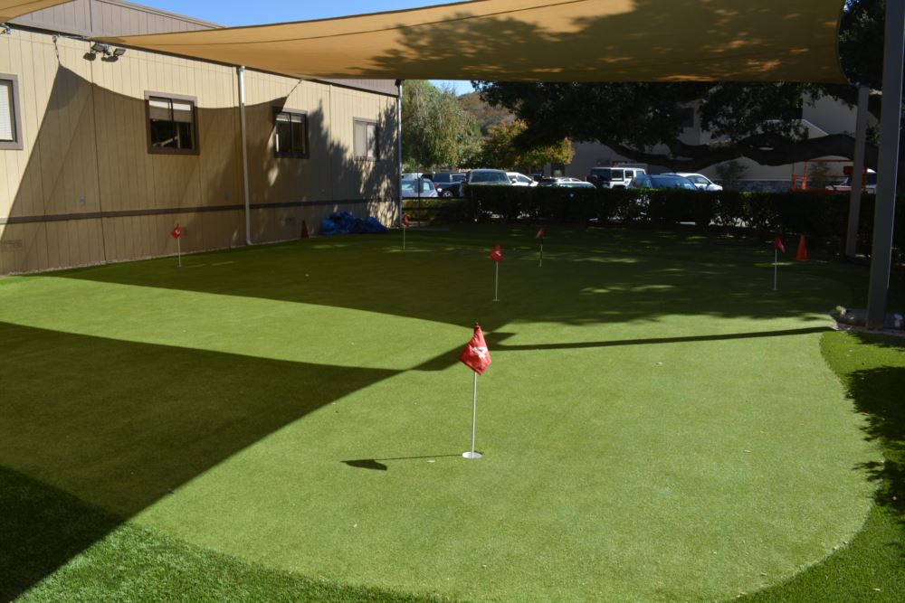 Multi-hole artificial putting green with red numbered flags and a fringe surround installed next to a commercial building under a shade sail