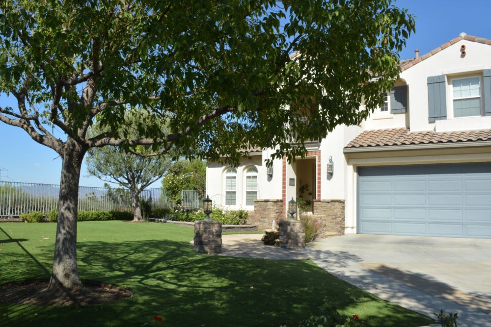 Artificial grass front lawn shaded by mature tree with stone entry pillars