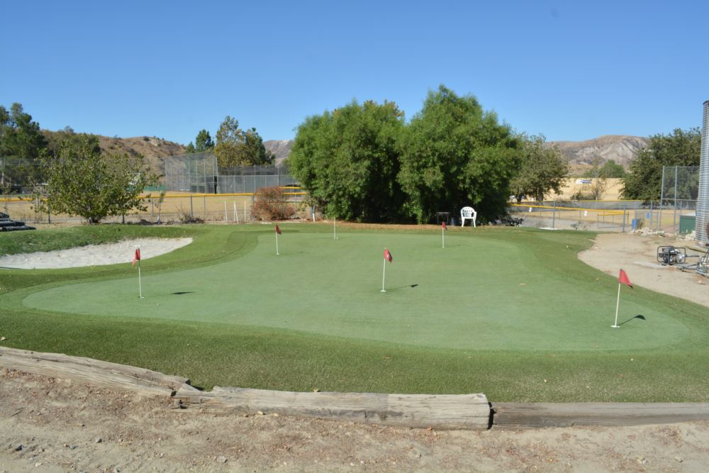 Large artificial putting green with five red flag pins, sand bunker, and contoured turf speed zones at a commercial sports complex beside a baseball field and desert hillside