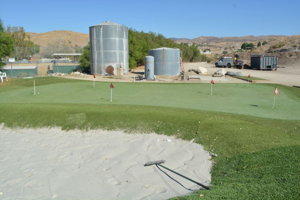 Artificial putting green with red flag pins and a sand bunker viewed from the bunker edge at a desert golf practice facility with water storage tanks nearby