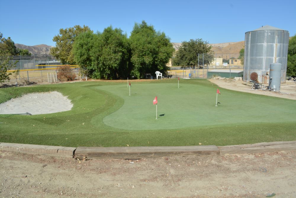 Commercial artificial putting green with red numbered flags, a sand bunker, and multi-speed turf zones at a rural sports complex with desert hills in the background
