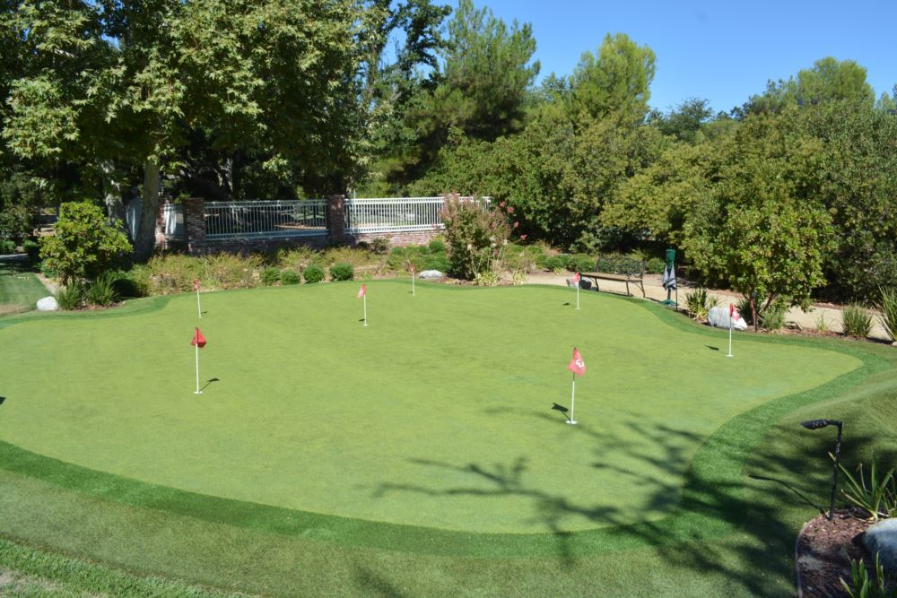 Custom backyard putting green with multiple red flag holes surrounded by mature trees and brick fence in Southern California