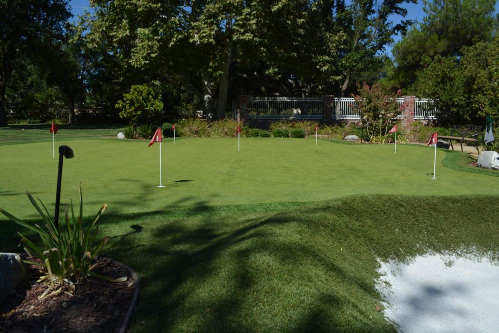 Large residential synthetic putting green with red flagsticks and sand bunker viewed from ground level with ornamental plantings and brick wall backdrop