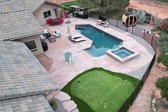 Aerial view of a luxury backyard featuring a synthetic grass installation putting green with multiple holes, complementing a resort-style pool, spa, stamped concrete patio, outdoor kitchen, and barrel sauna surrounded by Arizona red rock desert landscaping.