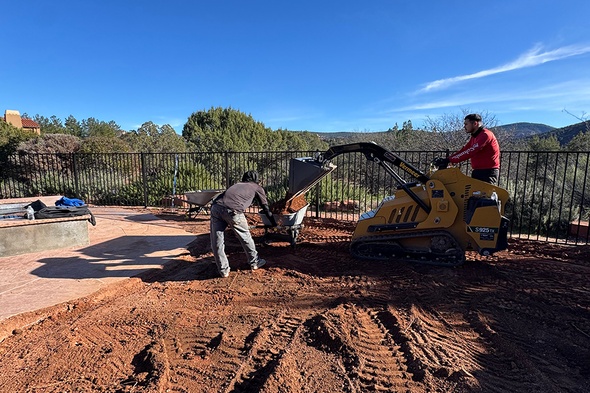 Professional crew using a compact track loader to grade and excavate red soil during a synthetic grass installation project in Sedona, Arizona, with a pool patio and mountain views in the background.
