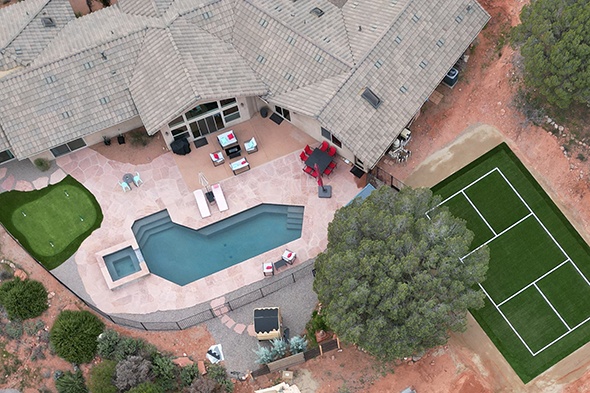 Drone view of a luxury Sedona, Arizona estate showcasing dual synthetic grass installations &mdash; a pickleball court with white boundary lines and a putting green &mdash; alongside a resort-style pool, spa, and stamped concrete patio surrounded by red rock desert terrain.