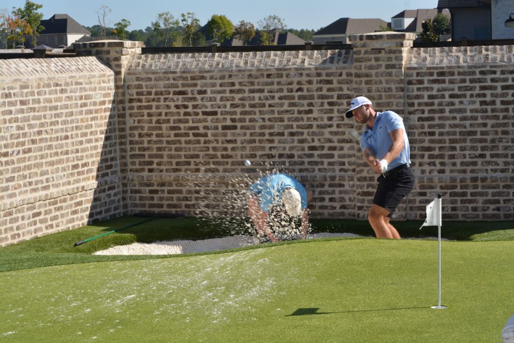 Sam Burns hitting a bunker shot on his custom Southwest Greens putting green, with sand splashing from the professional-grade practice bunker